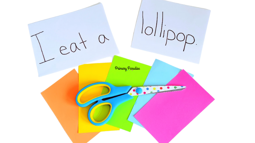 White background with the two pieces of white paper. On the first it says "I eat a" and the second says "lollipop. Underneath are 5 pieces of colorful paper, orange, yellow, green, blue, and pink, with scissors on top.