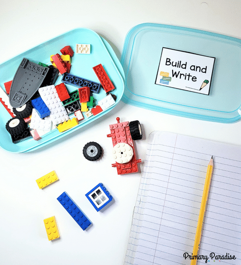 A box of legos with the lid to the right. The lid is labeled "build and write!" There are legos on the table and a blank notebook and pencils.