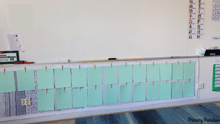 An image of a blank bulletin board under a white board with clothespins holding blank paper waiting for student work