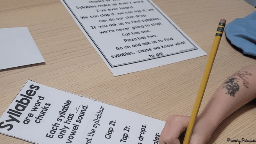 A child writing on a sheet that says "syllables are word chunks. Each syllable only has one vowel sound.