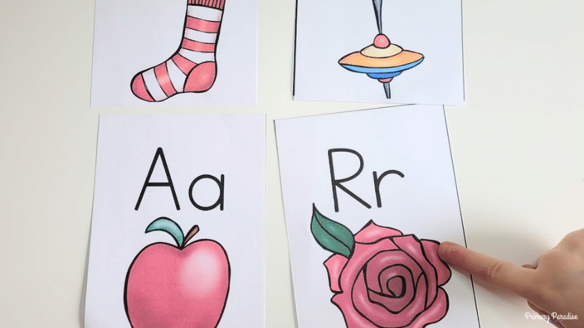 a child pointing to a flashcard with the letter r and a picture of a rose. To the left is a flashcard with the letter a and an apple