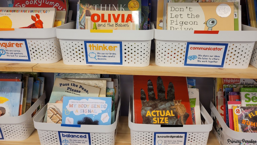 A picture of white bins with books on the. On each bin is a label with an IB learner profile characteristic and a definition of what it means.