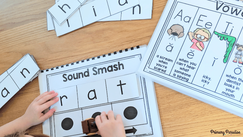 A child's hand moving a car across a phonics activity called "sound smash". In the top of the image are a stack of word cards and to the right is a vowel sound chart.