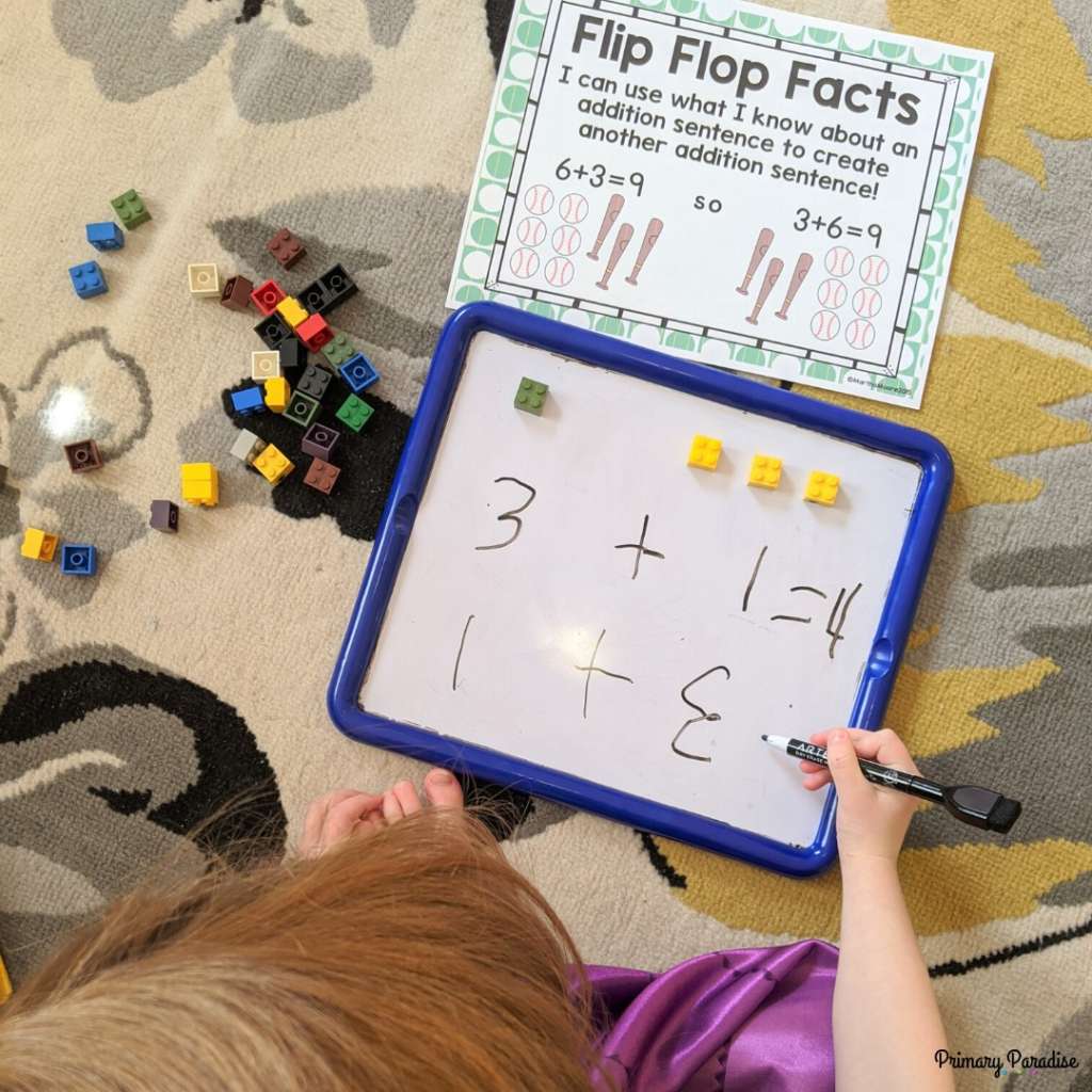 little girl writing on a white board. There is one green lego and 3 yellow legos, and she's writing 1+3 and 3+1