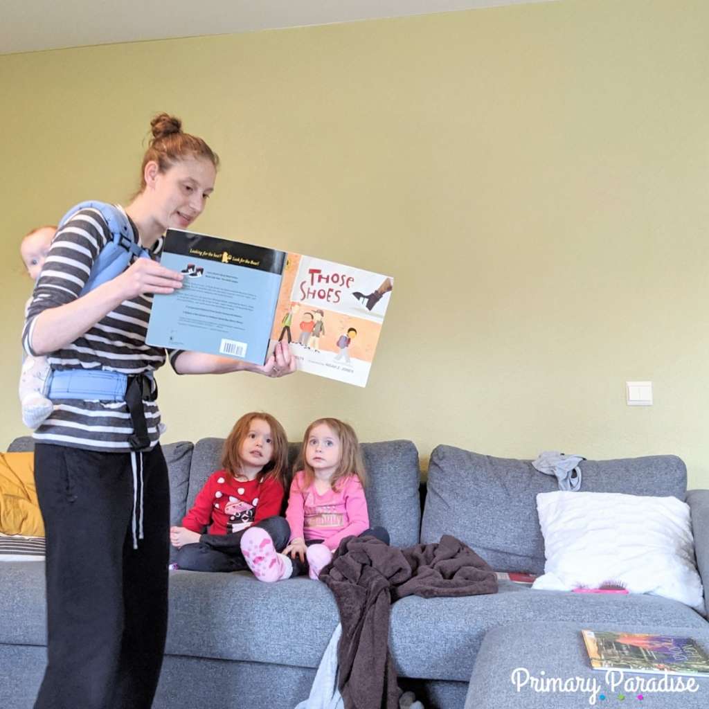 mom with baby on her back reading to two little girls on the couch. The book is "Those Shoes"